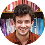 A young man with curly brown hair smiles brightly at the camera. He is in front of a shelf full of books organized by color.