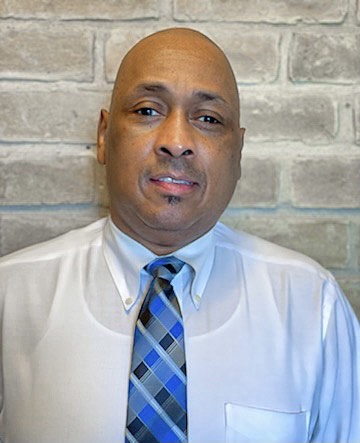 Head and shoulders of a smiling man in a white shirt and blue tie, against a tan brick wall.