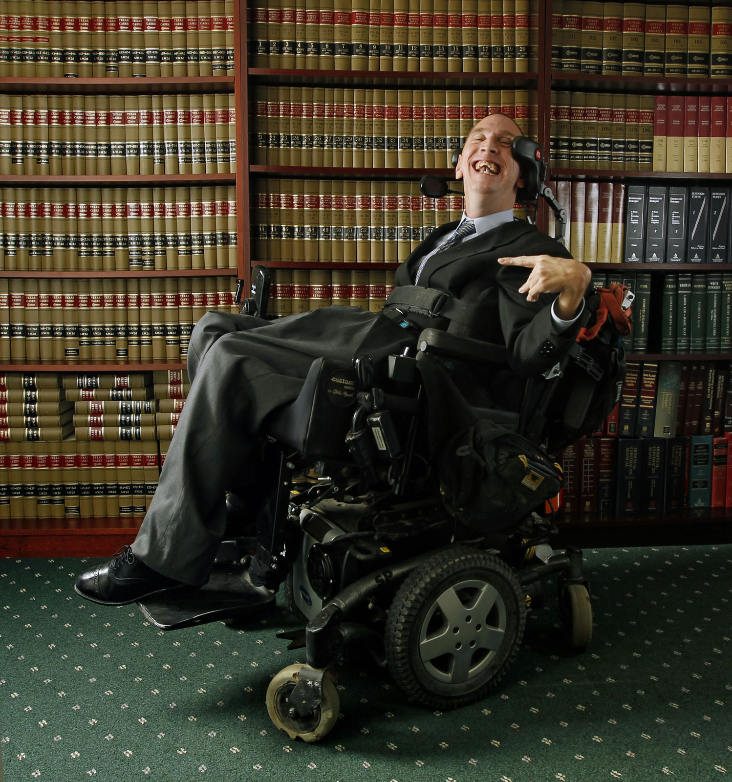 A man in a dark suit leans back in his power chair and smiles. Behind him is a full floor-to-ceiling bookcase.