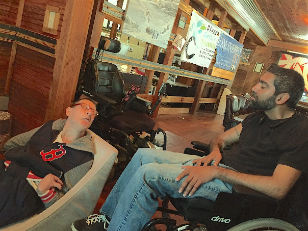 A woman seated in a legless chair on the floor twists to look up at a man behind her, seated in a wheelchair.