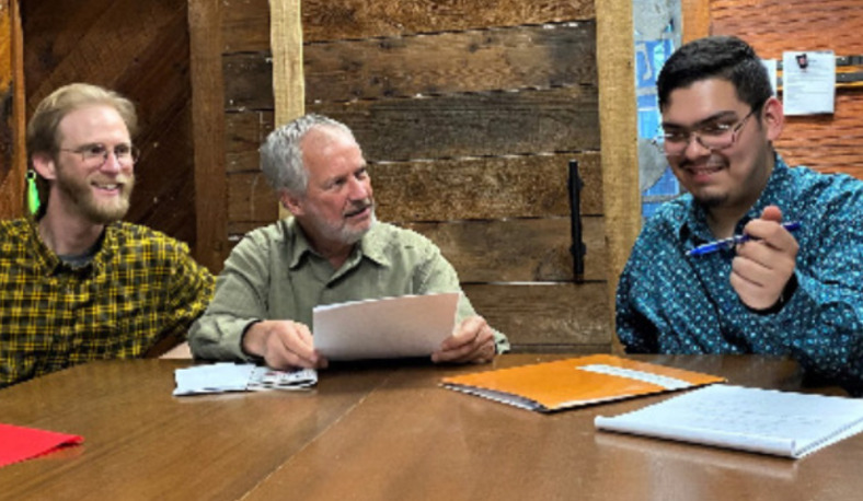 Three men sit at a wooden table. One gestures at a stack of papers with a pen while the other two attentively listen.