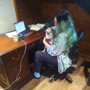 A woman with long green and black hair sits at a desk with a light tan Chihuahua in her lap.