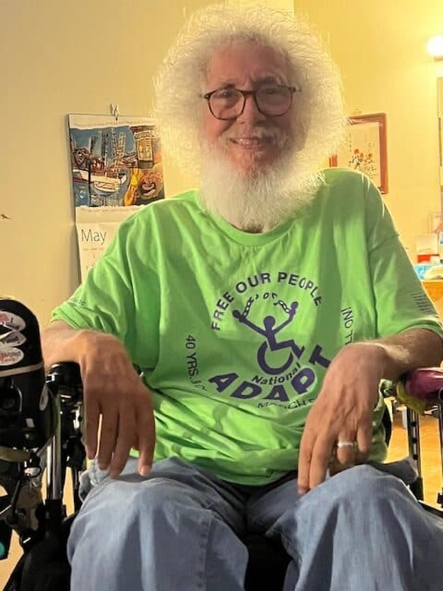 Activist Bob Kafka with white beard and hair wearing a green ADAPT t-shirt sitting in his wheelchair and smiling