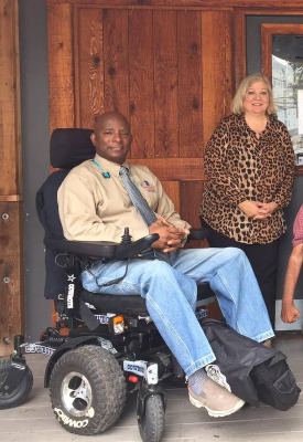 On a wooden porch, a man seated in a power chair and a woman standing next to him pose for the camera.