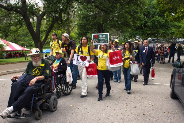 A man wearing a white, decorated straw hat and rolling a power chair looks at the camera as he leads a large group of marchers carrying signs.