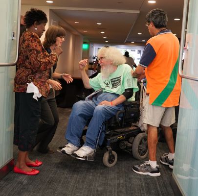 In a crowded walkway, a man with puffy white hair in a power chair is smiling and motioning to a young woman with glasses limply holding her hands out.