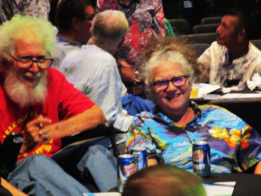 In a crowded room, a man with puffy white hair and beard and a woman wearing a Hawaiian shirt smile joyfully at the camera.