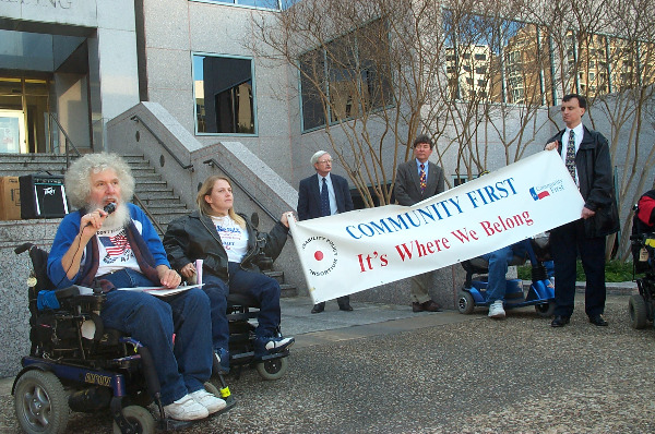 Outside the steps of a grey building, a man with puffy white hair and beard sits in a power chair and speaking into a microphone. Next to him, people are holding a banner that reads 
