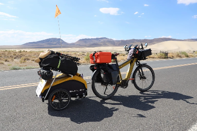E-bike loaded with bags and pulling a trailer is parked on the side of a sunny desert highway.