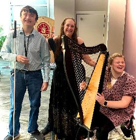 Three smiling people surround a small harp. A woman in the center puts her hand on the shoulder of a young man with a white cane. Her other hand is on the shoulder of a young seated woman with her hands at the harp-strings.