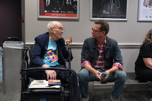 Two men, one with a walker, are seated and talking while gesturing with their hands.