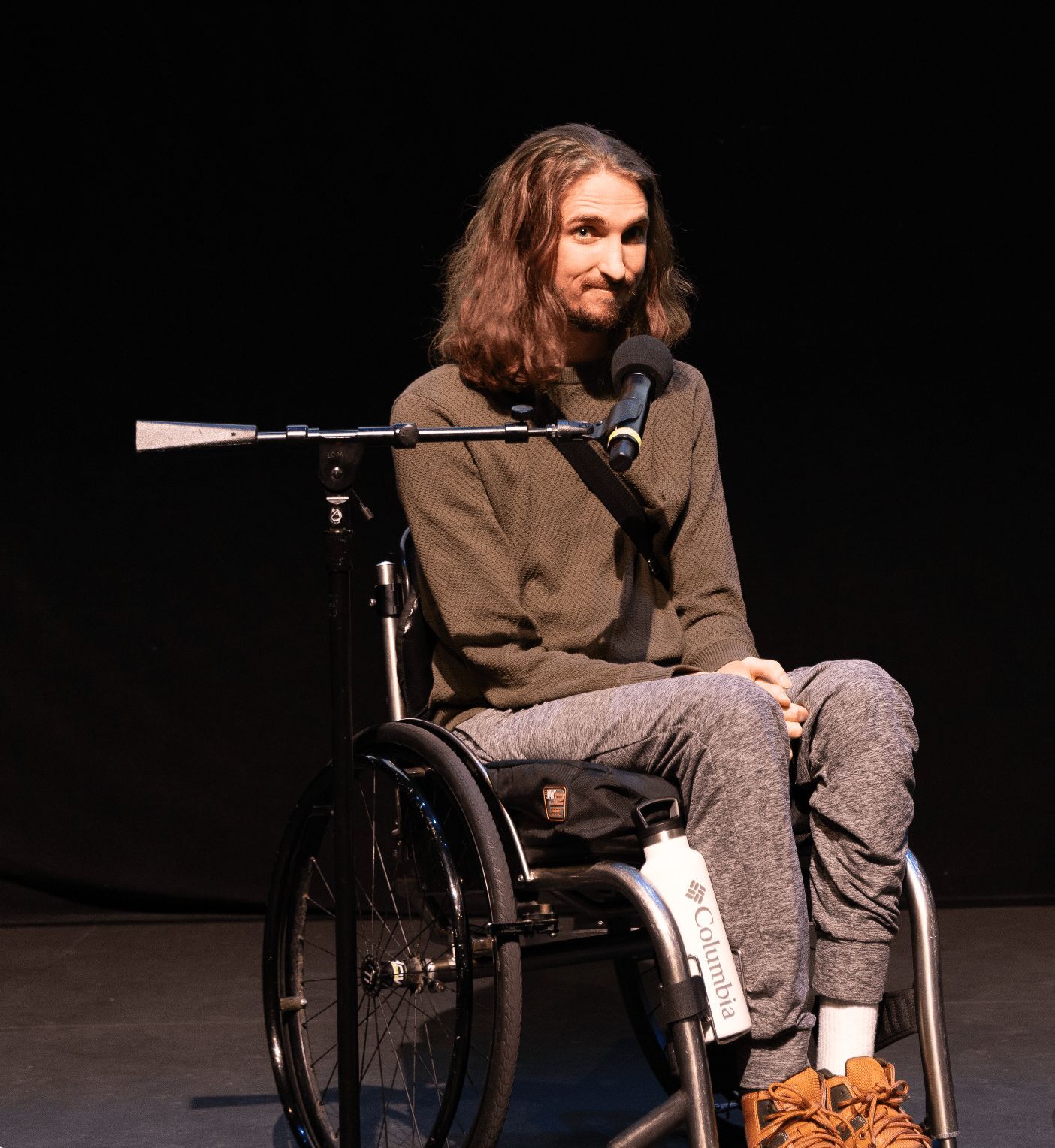 A white wheelchair user with long hair tells a joke into a microphone