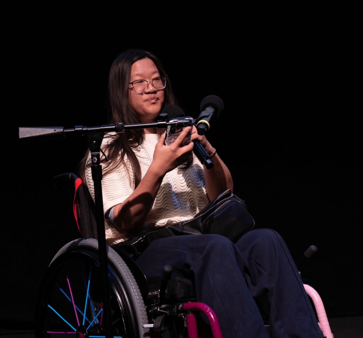 An Asian woman who uses a wheelchair reads off her phone into a microphone.