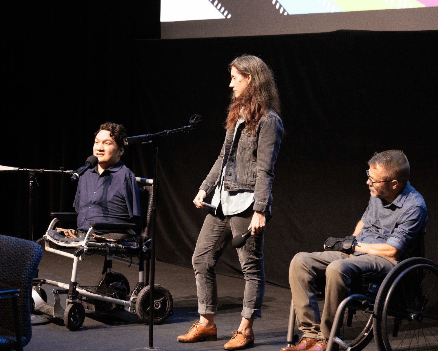 A white women and two men who use wheelchairs, one white one Latino, are onstage speaking into microphones.