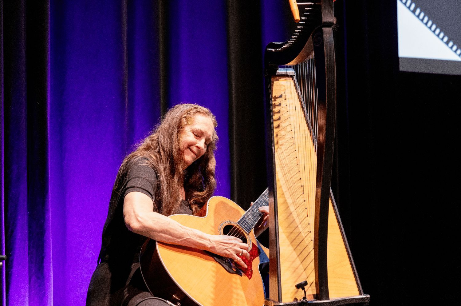 A white woman smiles and plays the acoustic guitar. A harp and the corner of the film screen are in the background.