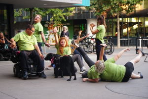 Across a paved area, a group of people in matching line green shirts are in various positions: sitting or lying on the ground, sitting in a wheelchair, standing. Arms and legs are held out at various angles.